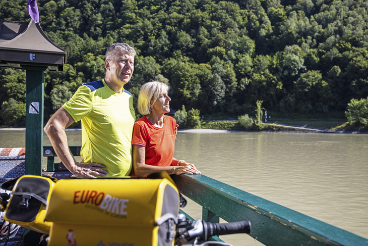 Zwei Radfahrer auf einer Fähre auf der Donau mit Fahrrädern im Vordergrund.