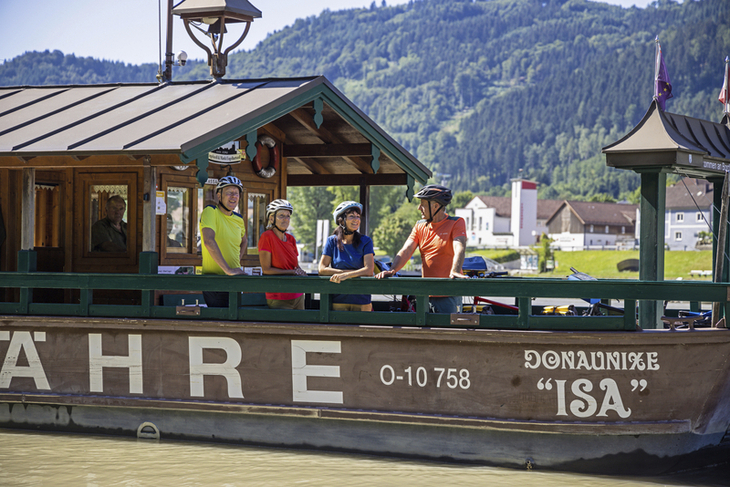 Radfahrer auf einer Fähre am Donau-Radweg, Berge im Hintergrund.