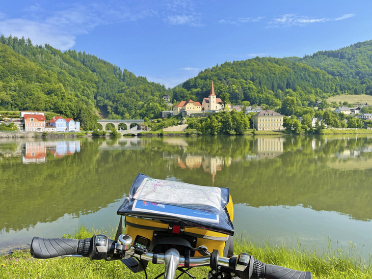 Blick auf eine Stadt am Fluss mit Brücke und bewaldeten Hügeln, Fahrradlenker im Vordergrund.