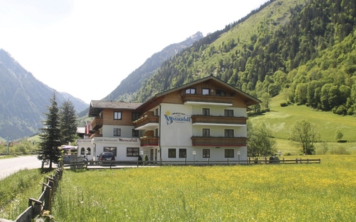 Hotelgebäude in bergiger Landschaft mit Wiesen und Wald im Hintergrund.