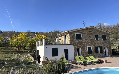 Steinhaus mit Terrasse, umgeben von Garten und Pool bei sonnigem Wetter.