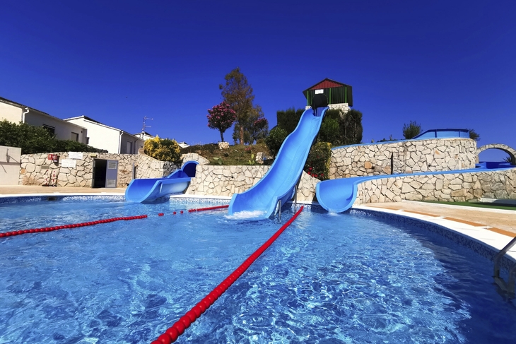Blauer Swimmingpool mit drei Wasserrutschen neben Steingarten und Gebäuden unter klarem Himmel.