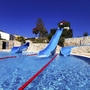 Blauer Swimmingpool mit drei Wasserrutschen neben Steingarten und Gebäuden unter klarem Himmel.