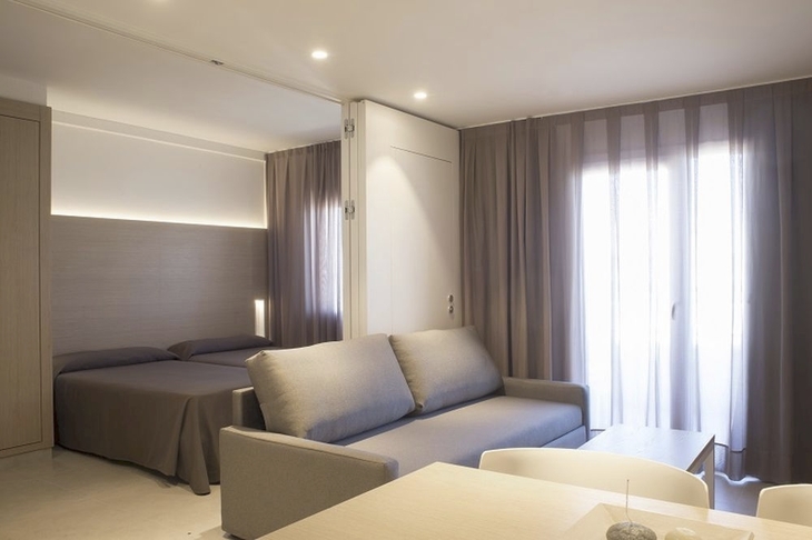 Moderne Wohnung mit Sofa, Esstisch, Doppelbett und großen Fenstern mit Vorhängen.