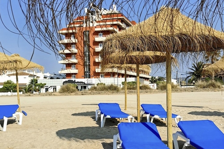 Sonnenliegen unter Strohdächern am Strand vor einem Hotel.