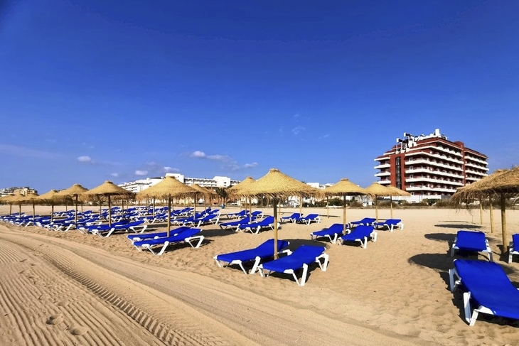 Strand mit blauen Liegen und Strohhütten vor einem Hotel.
