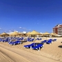 Strand mit blauen Liegen und Strohhütten vor einem Hotel.