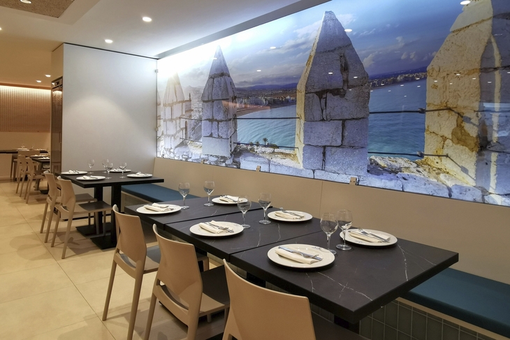 Restaurant mit gedeckten Tischen und Wandfoto von Meer und Mauer.