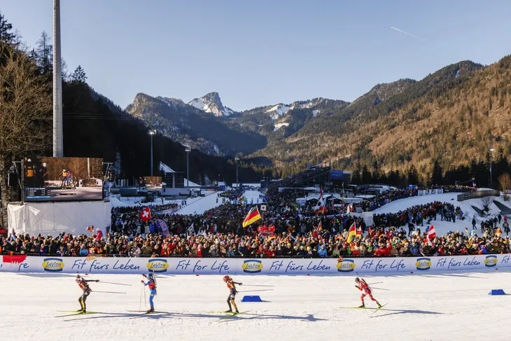 Biathleten in der Chiemgau Arena bei Staffellauf, zahlreiche Zuschauer, Berge im Hintergrund.
