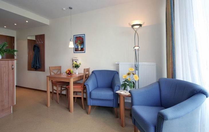 Wohnzimmer mit blauen Sesseln, Esstisch, Blumen und heller Beleuchtung.