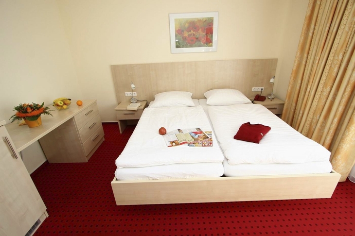 Hell eingerichtetes Hotelzimmer mit Doppelbett und roten Teppichboden.
