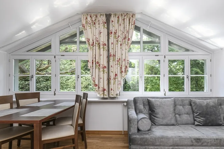 Wohnraum mit großem Fenster, Vorhängen, Couch und Esstisch.