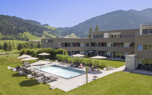 Gebäude mit Pool, Sonnenschirmen und grüner Hügellandschaft im Hintergrund.