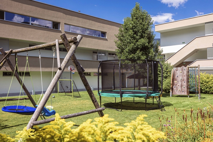 Spielplatz mit Schaukel, Trampolin und Kletterwand im Freien bei sonnigem Wetter.