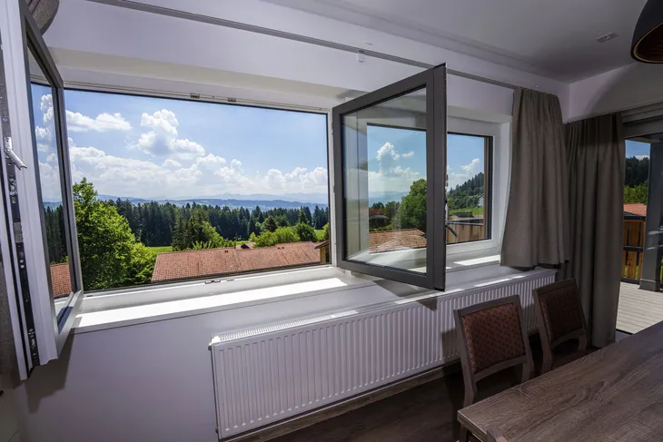 Blick aus einem geöffneten Fenster auf eine Waldlandschaft und blauen Himmel.