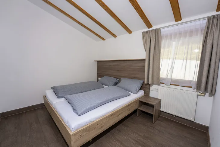 Kleines Schlafzimmer mit Doppelbett, Holzbalkendecke und Fenster mit Vorhang.