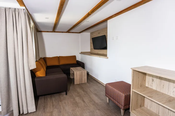 Wohnraum mit Ecksofa, Fernseher an der Wand, Holzboden, Bücherregal und kleinem Tisch.
