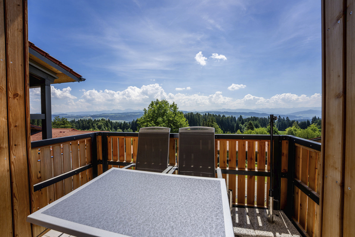 Balkon mit Tisch und Stühlen, Blick auf Wald und Berge unter blauem Himmel.