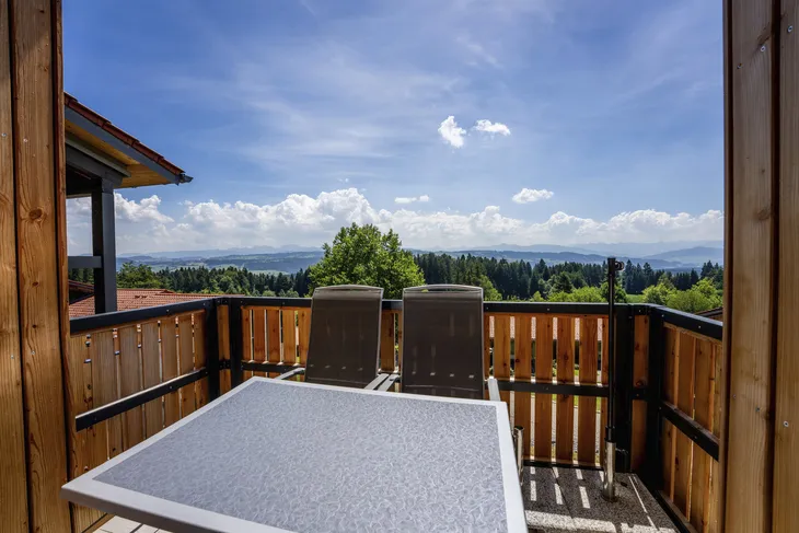 Balkon mit Tisch und Stühlen, Blick auf Wald und Berge unter blauem Himmel.