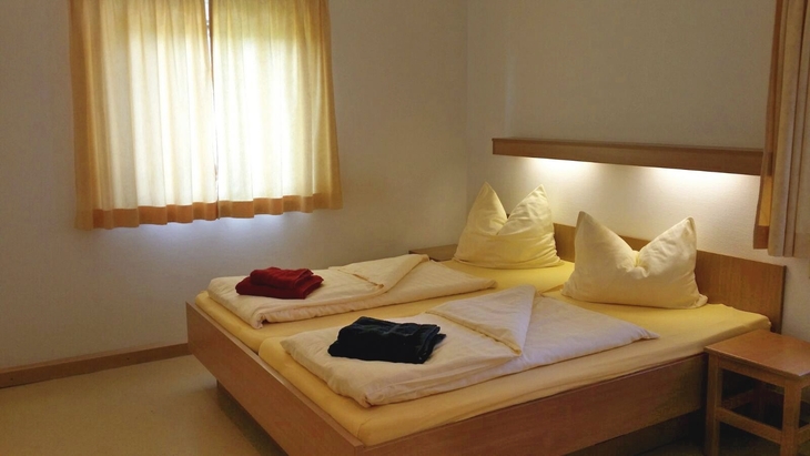 Gemütliches Schlafzimmer in der Ferienanlage Scheidegg, mit Doppelbett, Bettwäsche, Handtüchern und Nachttisch, sowie einem Fenster mit gelben Vorhängen.