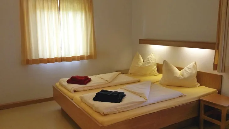Gemütliches Schlafzimmer in der Ferienanlage Scheidegg, mit Doppelbett, Bettwäsche, Handtüchern und Nachttisch, sowie einem Fenster mit gelben Vorhängen.