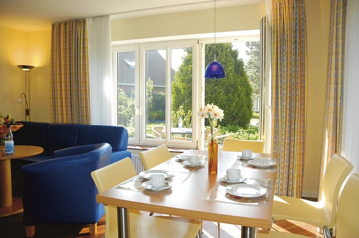Wohnbereich mit Esstisch, blauen Sesseln und Blick auf Garten in Ferienanlage Harlesiel.