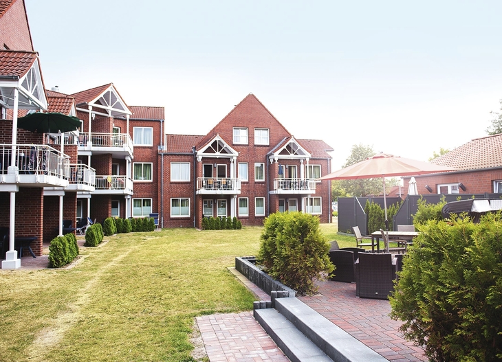 Außenaufnahme der Ferienanlage Büsum, bestehend aus aneinandergebauten roten Klinkergebäude mit spitzen Dächern und weißen Balkonen, vor sonnigem Himmel. Rechts im Bild eine Terrasse mit Sitzmöbeln und zwischen den Häusern und der Terrasse ein breiter Grünstreifen. 