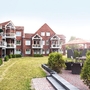 Außenaufnahme der Ferienanlage Büsum, bestehend aus aneinandergebauten roten Klinkergebäude mit spitzen Dächern und weißen Balkonen, vor sonnigem Himmel. Rechts im Bild eine Terrasse mit Sitzmöbeln und zwischen den Häusern und der Terrasse ein breiter Grünstreifen. 