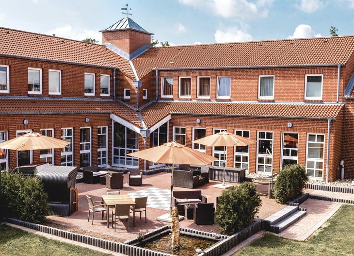 Rotes rechtwinkliges Backsteingebäude der Ferienanlage Büsum mit Terrasse, Sitzmöbeln und Sonnenschirmen bei sonnigem Wetter.