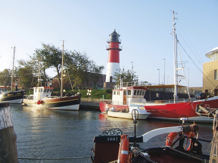 Boote im Hafen von Büsum vor einem Leuchtturm bei klarem Himmel.