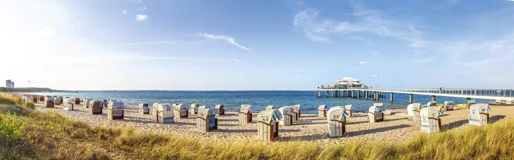 Strandkörbe am Timmendorfer Strand mit Seebrücke im Hintergrund und Seegras im Vordergrund, bei leichtbewölktem Himmel.