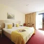 Helles Zimmer mit rotem Teppichboden im Haus Oldenburg in der Ferienanlage Timmendorfer Strand. Doppelbett mit gelber Bettwäsche, Nachtischen und zwei Stühlen mit kleinem Tisch, sowie einem Schreibtisch und Balkontür mit großem gelb-rotem Vorhang.