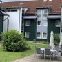 Mehrstöckiges Gebäude mit Terrasse mit Tischen, Stühlen und Sonnenschirmen und grünem Busch auf einer Wiese.