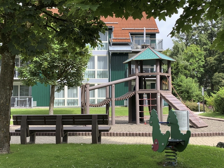 Spielplatz mit Spielturm und Wipptier sowie einer Holzbank vor einem Gebäude der Ferienanlage Braunlage.