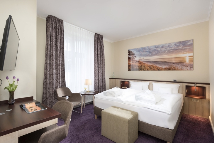 Hotelzimmer mit Doppelbett, Schreibtisch, zwei Stühlen und Landschaftsbild an der Wand.