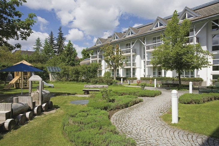 Gebäude mit Garten, Spielplatz und gepflastertem Weg bei wolkigem Himmel.