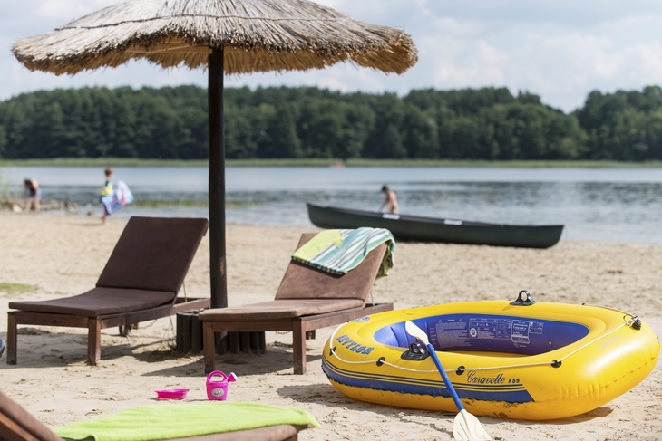 Strand mit Liegen, Sonnenschirm und gelbem aufblasbarem Paddelboot am Seeufer.