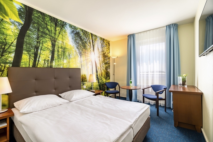 Helles Hotelzimmer mit Doppelbett, Waldtapete und blauen Vorhängen.