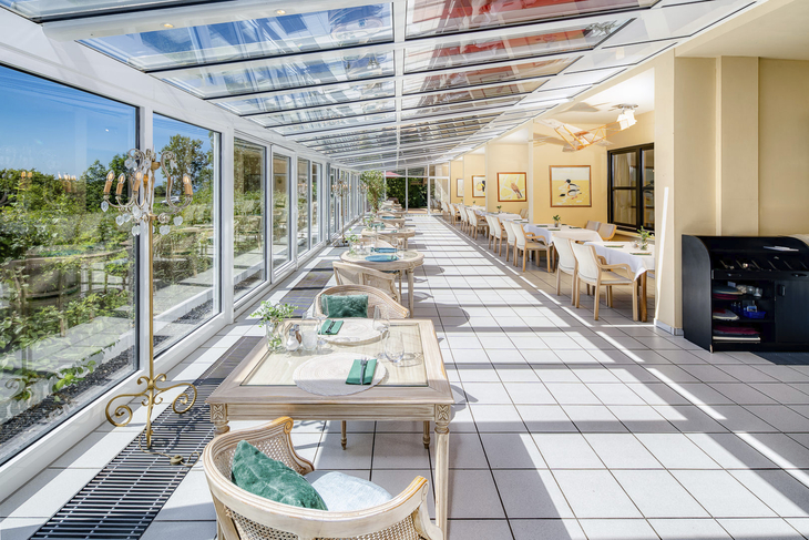 Heller Wintergarten eines Hotels mit gedeckten Tischen und Blick ins Grüne.