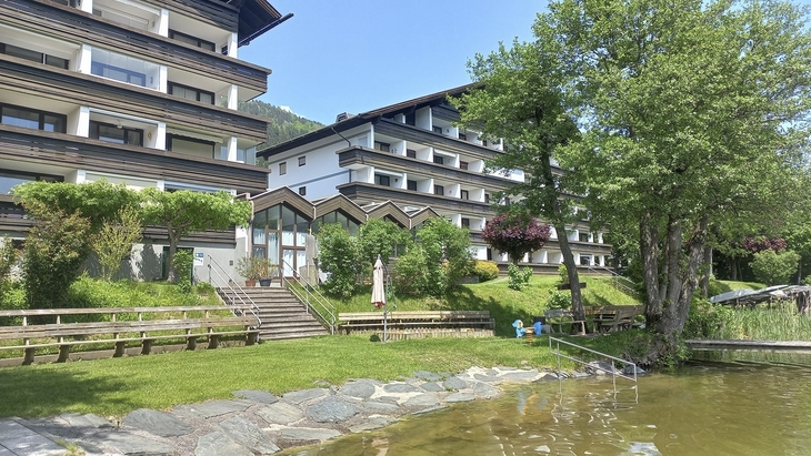Appartementanlage am See mit Garten und Bäumen an einem sonnigen Tag.