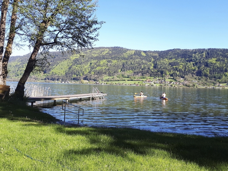 Zwei Personen paddeln auf einem See neben einem Steg, umgeben von Hügeln und Bäumen.