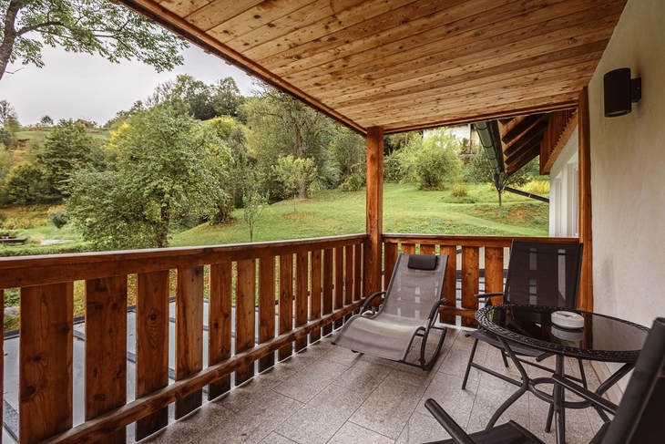 Überdachte Terrasse mit Holzgeländer und Gartenmöbeln vor bewaldeter Landschaft.