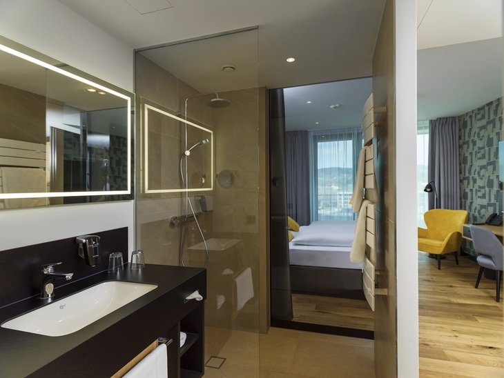 Modernes Hotelzimmer mit Glasdusche, Doppelbett und gelbem Sessel.
