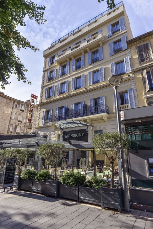 Fassade des Hotel Monsigny in Nizza mit Café-Terrasse und Straßenszene.