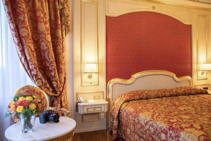 Luxuriöses Hotelzimmer mit Blumen, Vorhängen und Bett in Rot-Gold-Tönen.