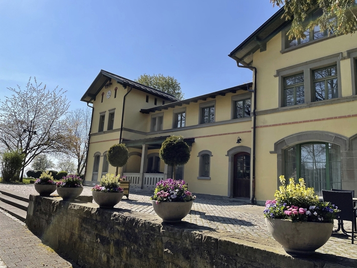 Gelbes Gebäude mit bepflanzten Blumenkübeln im Vordergrund bei sonnigem Wetter.