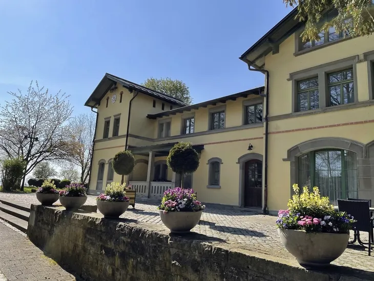 Gelbes Gebäude mit bepflanzten Blumenkübeln im Vordergrund bei sonnigem Wetter.