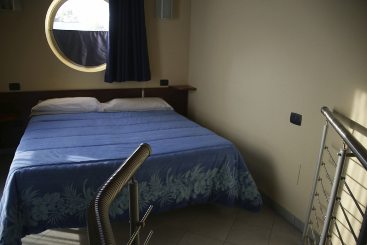 Hotelzimmer mit Bett, blauem Bettüberwurf und rundem Fenster.