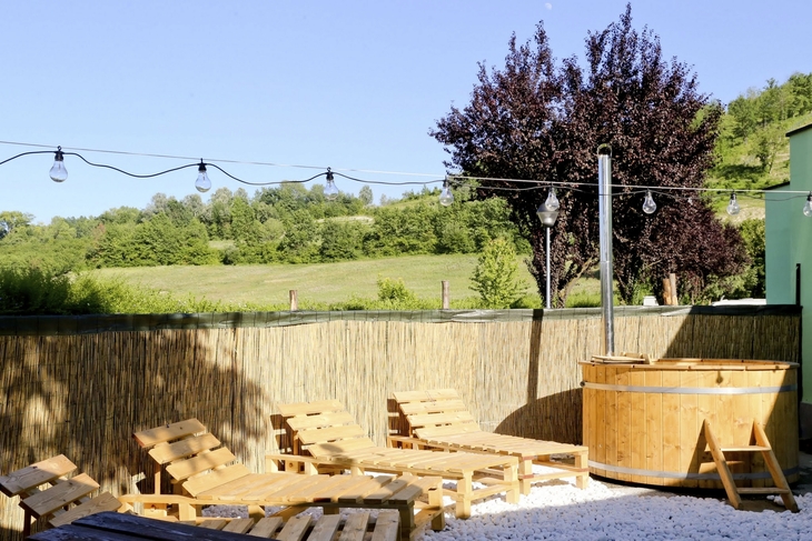 Außenbereich mit Holzliegen, Hecke und Baum vor Hügelkulisse.