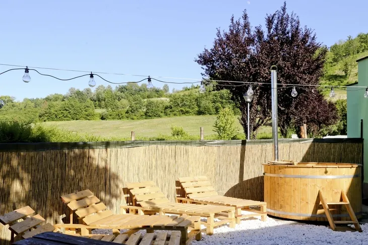 Außenbereich mit Holzliegen, Hecke und Baum vor Hügelkulisse.